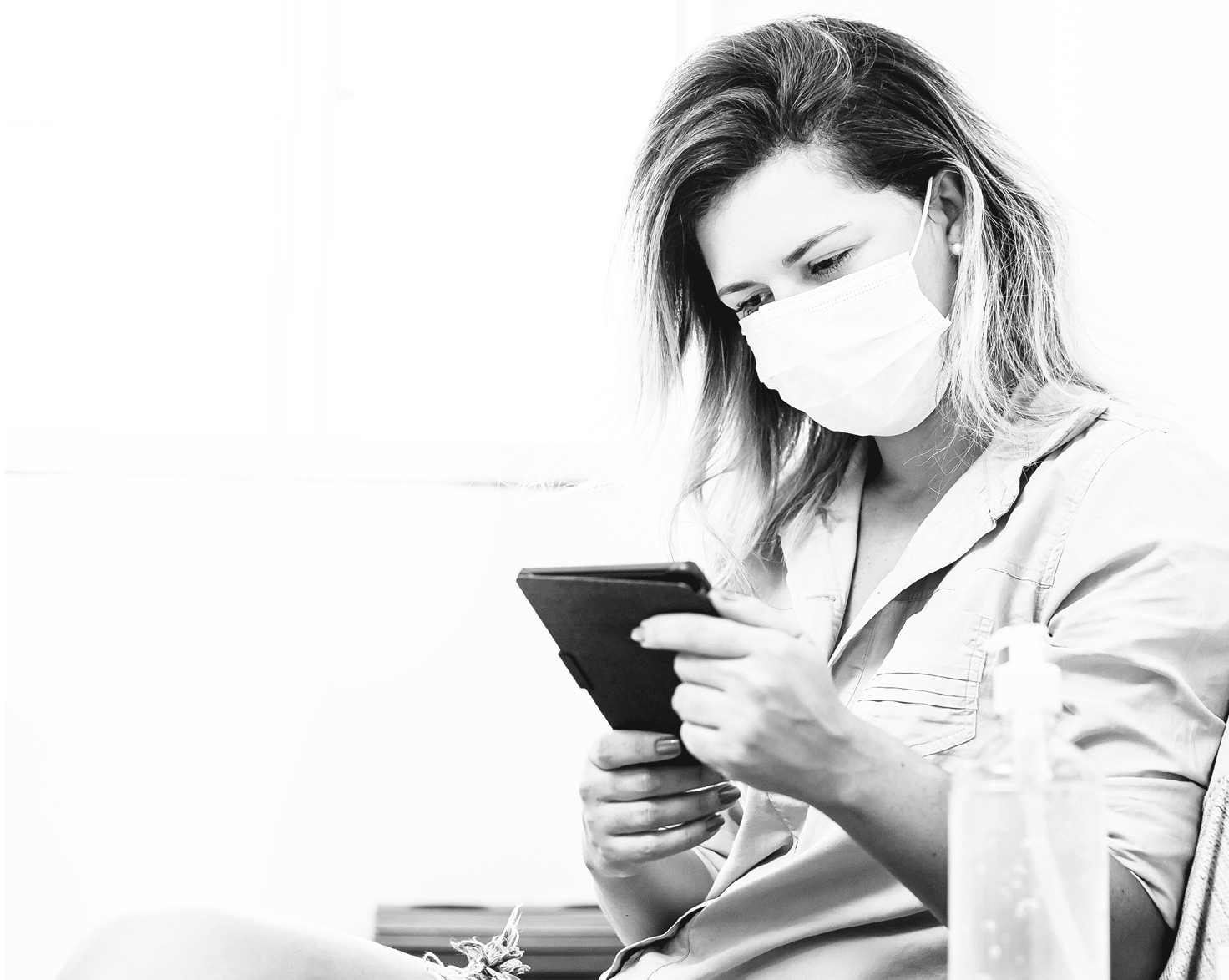 Coronavirus  Covid-19  Woman in quarantine wearing protective mask reading e-book in the living room  Bottle of alcohol gel in the foreground 